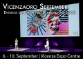 Zwei Personen auf der Bühne des Vicenza Expo Centre während der Präsentation bei Vicenzaoro September. Im Hintergrund ist eine große Leinwand zu sehen, die Schmuckstücke und eine Hand mit Ringen zeigt. Text auf dem Bild: 'Vicenzaoro September - Entdecke die Zukunft des Schmucks und Luxus! 6. - 10. September | Vicenza Expo Centre'.