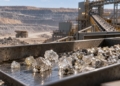 Rohdiamanten auf einem Förderband vor einer Diamantmine mit Aufbereitungsanlage und Muldenkipper im Hintergrund