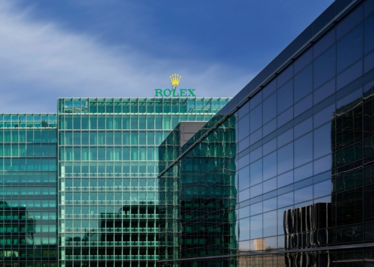 Glasfassade der Rolex-Zentrale mit Rolex-Schriftzug und Krone auf dem Dach vor blauem Himmel